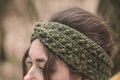 A close-up of a woman wearing a khaki green chunky crochet headband outdoors with a blurred natural background.