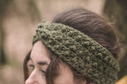 A close-up of a woman wearing a khaki green chunky crochet headband outdoors with a blurred natural background.