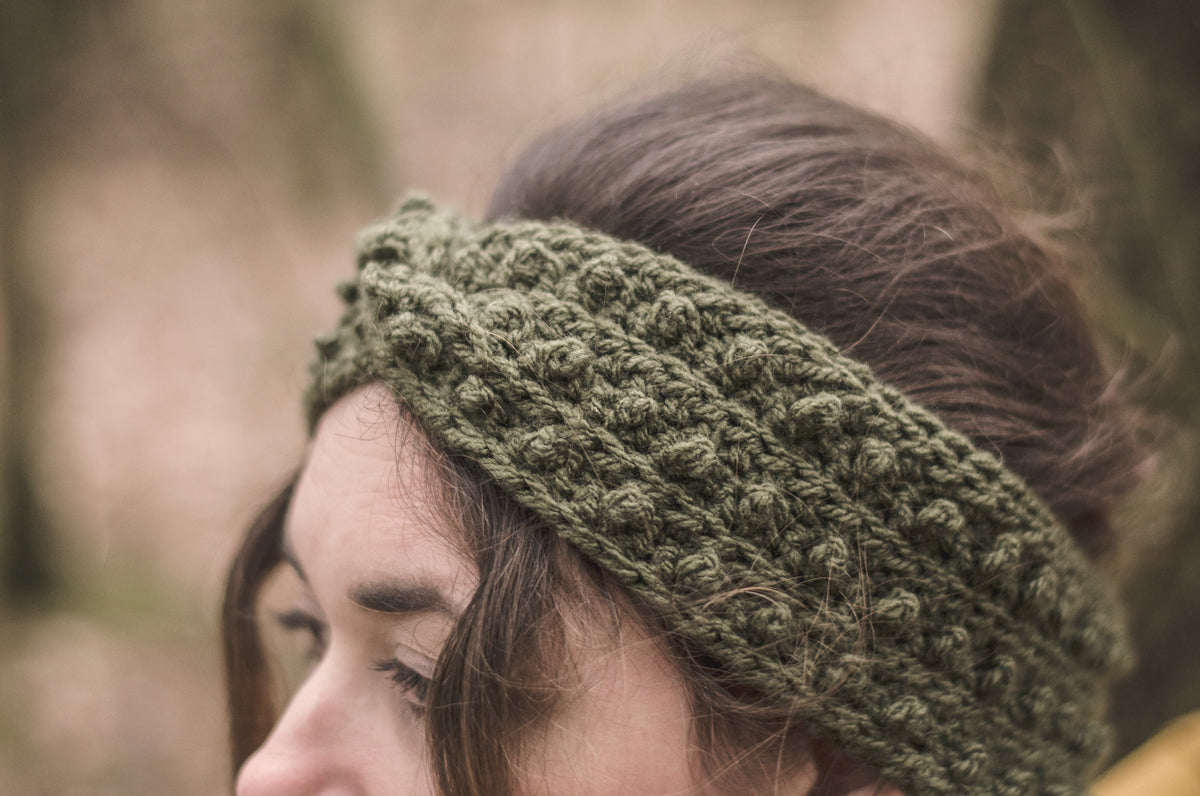 A close-up of a woman wearing a khaki green chunky crochet headband outdoors with a blurred natural background.