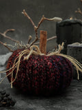 A crocheted decorative pumpkin in black and jewel tone variegated yarn, featuring a cinnamon stick stem and rustic jute twine bows, styled on a dark textured surface with dried branches and candles.