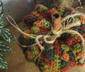A close-up of a crocheted decorative pumpkin in autumn colours, featuring textured stitches in orange, green, and red-brown, with a cinnamon stick stem and jute twine bow, set against a neutral background with houseplant leaves on the left.
