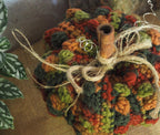 A close-up of a crocheted decorative pumpkin in autumn colours, featuring textured stitches in orange, green, and red-brown, with a cinnamon stick stem and jute twine bow, set against a neutral background with houseplant leaves on the left.