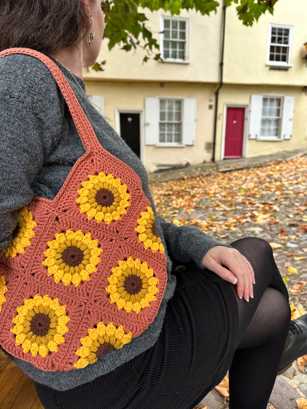 A woman sitting outdoors on a bench wearing a crocheted shoulder bag with a sunflower pattern. The bag features alternating yellow and mustard sunflower motifs with dark brown centres, set against a background of a yellow building and fallen autumn leaves.
