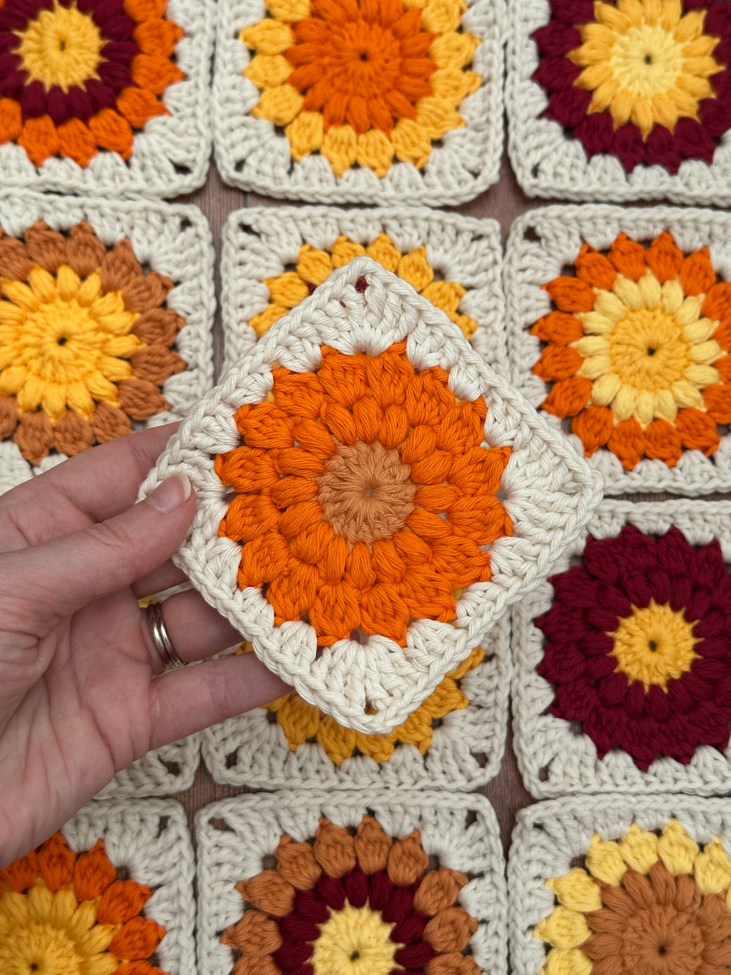 Hand holding a crochet granny square with sunflower pattern above a background of more crocheted granny squares.