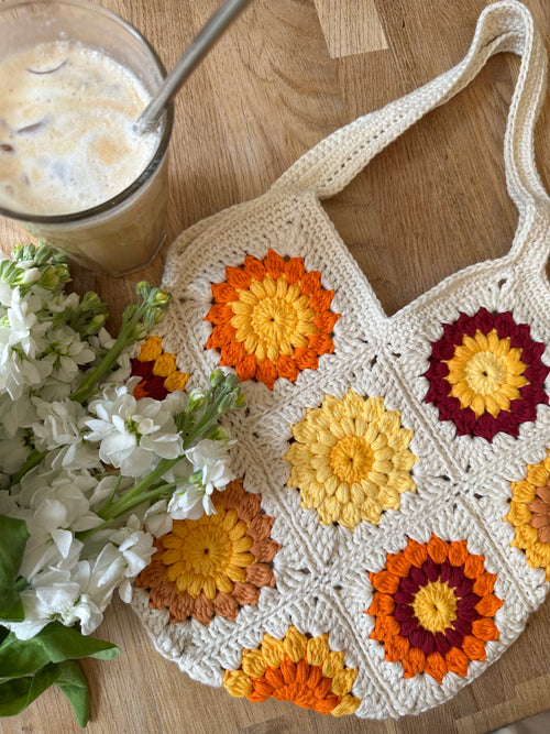 Crocheted bag with floral motifs on a wooden table with a glass of iced coffee and fresh white flowers.