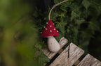 A small crocheted mushroom ornament with a jute hanging string, propped on a rustic wooden bench, surrounded by green ivy foliage.