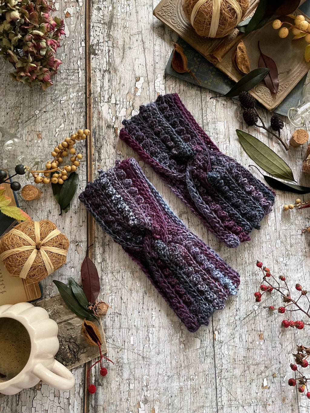 Two chunky crochet headwarmers in a variegated purple, blue, and gray yarn, styled in a flat-lay on a rustic white wooden table surrounded by autumn-themed props.