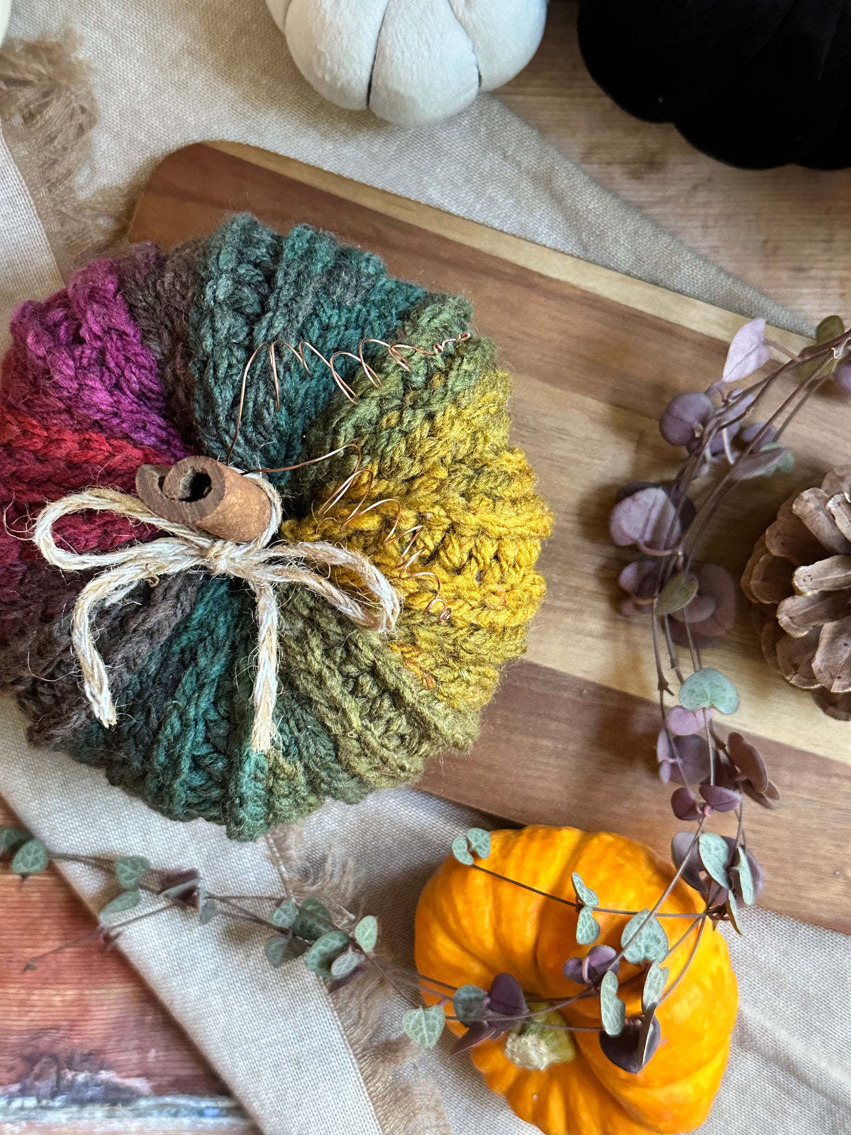 A crocheted decorative pumpkin  in a rainbow autumn gradient, featuring shades of green, yellow, blue and pink/purple, tied with a rustic jute twine bow and a cinnamon stick. The bundle is styled on a wooden cutting board with autumn-themed props including a small orange pumpkin, pinecones, houseplant vines, and a white pumpkin.