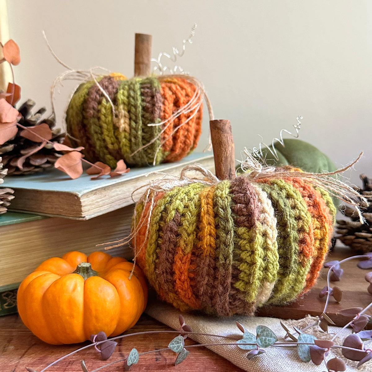 Two crocheted autumn pumpkins with cinnamon stick stems and jute twine accents, styled on vintage books with a real orange pumpkin and pinecones on a rustic wooden table.