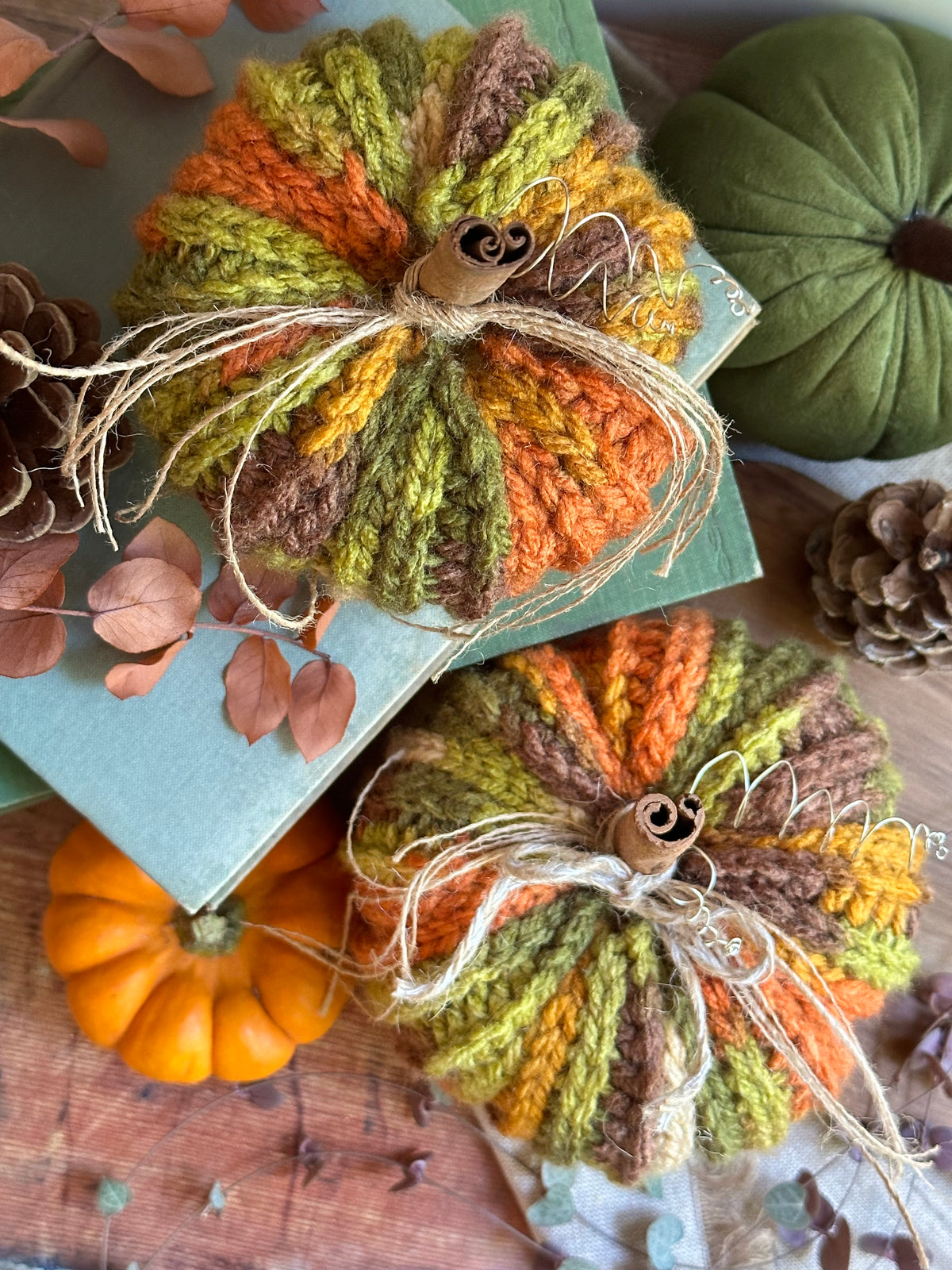 Two crocheted yarn pumpkins in autumn colours of orange, olive green, and brown, decorated with twine and cinnamon sticks, styled on a wooden surface with small pumpkins and pinecones.