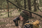 A woman hiking in a forest, wearing a khaki green crocheted headwarmer and matching fingerless gloves, drinking from a black insulated tumbler with a patterned sleeve. She has a yellow hiking backpack on her shoulder.