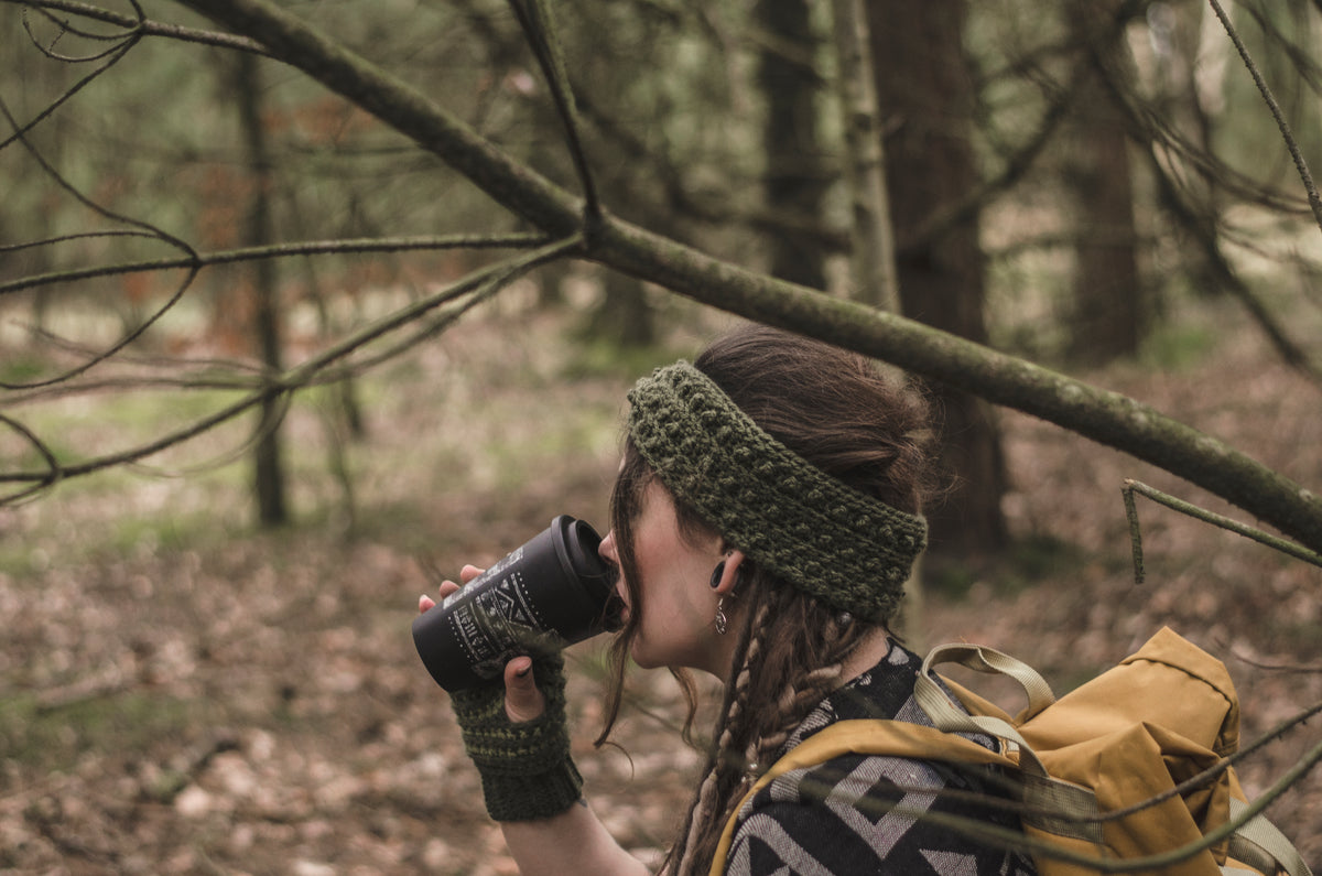 A woman hiking in a forest, wearing a khaki green crocheted headwarmer and matching fingerless gloves, drinking from a black insulated tumbler with a patterned sleeve. She has a yellow hiking backpack on her shoulder.