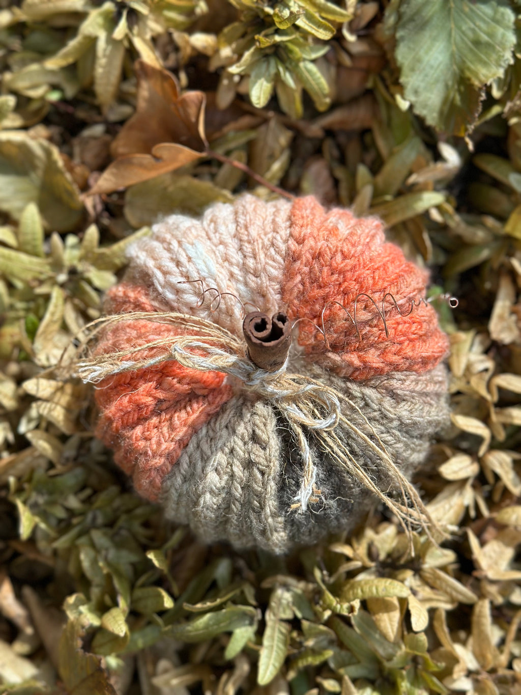 A crocheted decorative pumpkin ornament in shades of orange/pink, beige, and grey, with a cinnamon stick stem and twine details, resting on dry leaves and foliage.