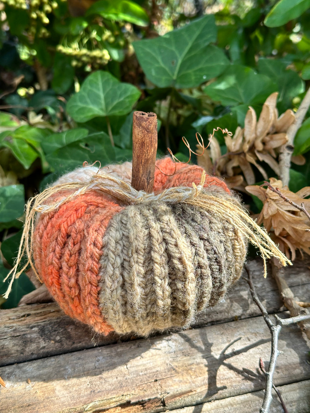 A crocheted decorative pumpkin with a rustic cinnamon stick stem, featuring colour-pooled sections of orange/pink, beige, and cream yarn, resting on a weathered wooden surface with green ivy in the background.