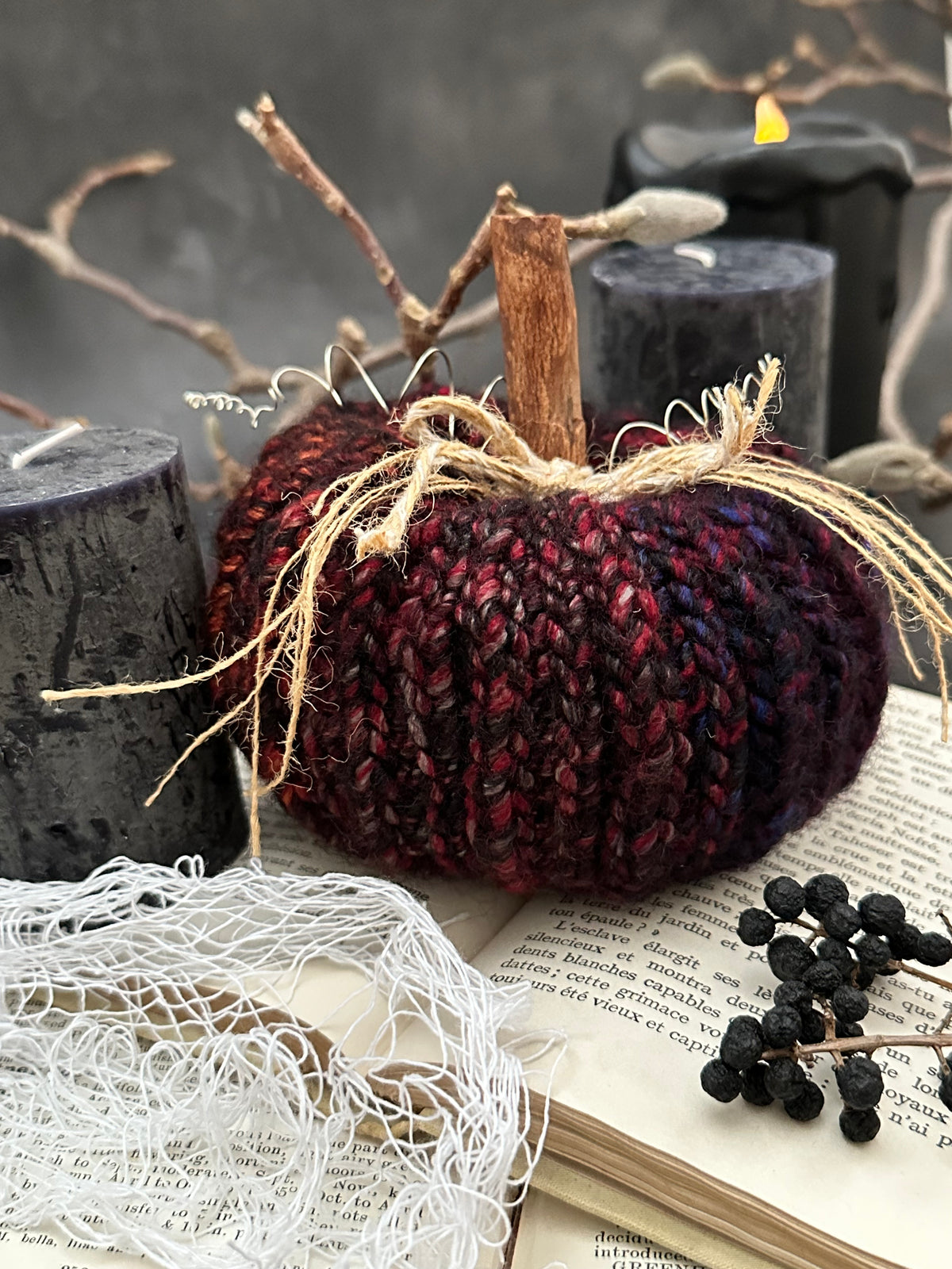 A crocheted decorative pumpkin in black and jewel tone variegated yarn, styled on an open vintage book with black candles, white cobweb fabric, and dried branches.