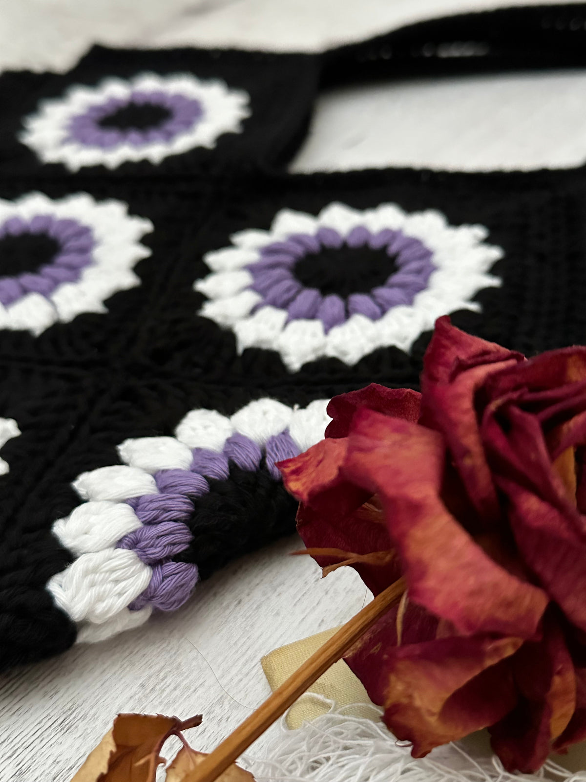 A black crocheted market bag with granny square floral motifs in black, purple and white, laid flat on a white wood surface with a dried rose.