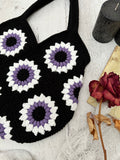 A black crocheted market bag with granny square floral motifs in black, purple and white, laid flat on a white wood surface with a dried rose, vintage book, and black pillar candles.