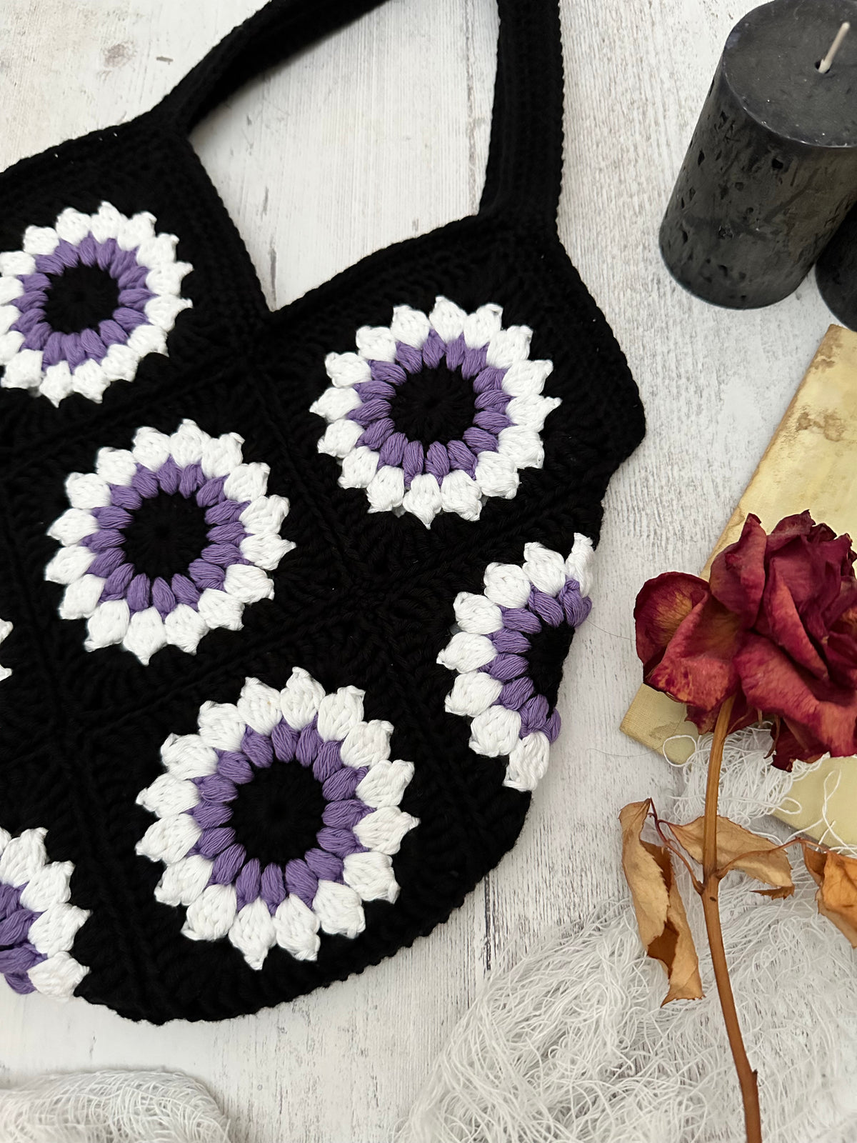 A black crocheted market bag with granny square floral motifs in black, purple and white, laid flat on a white wood surface with a dried rose, vintage book, and black pillar candles.