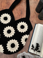 A black crocheted tote bag with large cream-coloured sunflower motifs, laid flat on a wooden surface next to an open book and dried flowers.