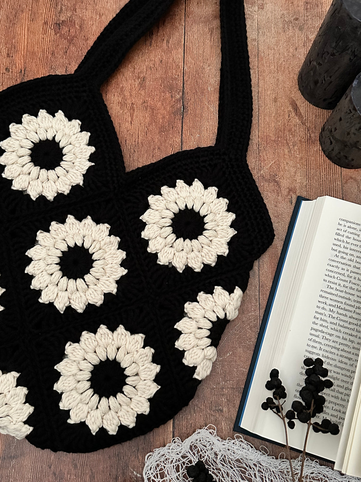 A black crocheted tote bag with large cream-coloured sunflower motifs, laid flat on a wooden surface next to an open book and dried flowers.