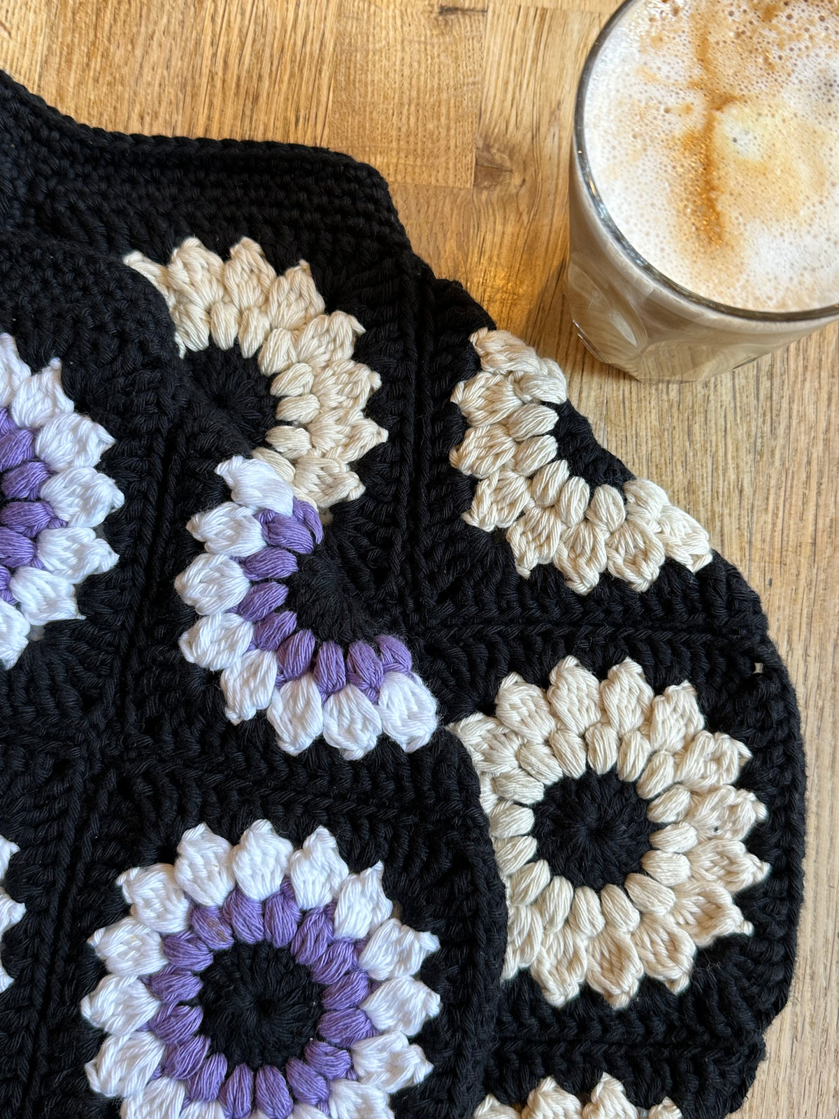 A close-up of two black themed crocheted bags with granny square floral motifs in black/purple/white and black/neutral colours, laid on a wooden table next to a glass of iced coffee.