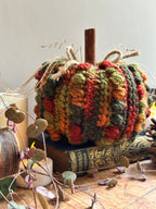 A handmade, chunky crocheted decorative pumpkin in autumn colours of orange, green, and red, styled on vintage books with houseplant vines and candles.