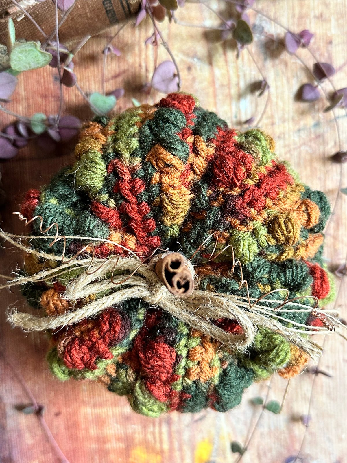 A handmade crocheted pumpkin ornament in autumn colours of green, orange, and red, tied with twine and a cinnamon stick, set against a rustic wood background with houseplant vines.
