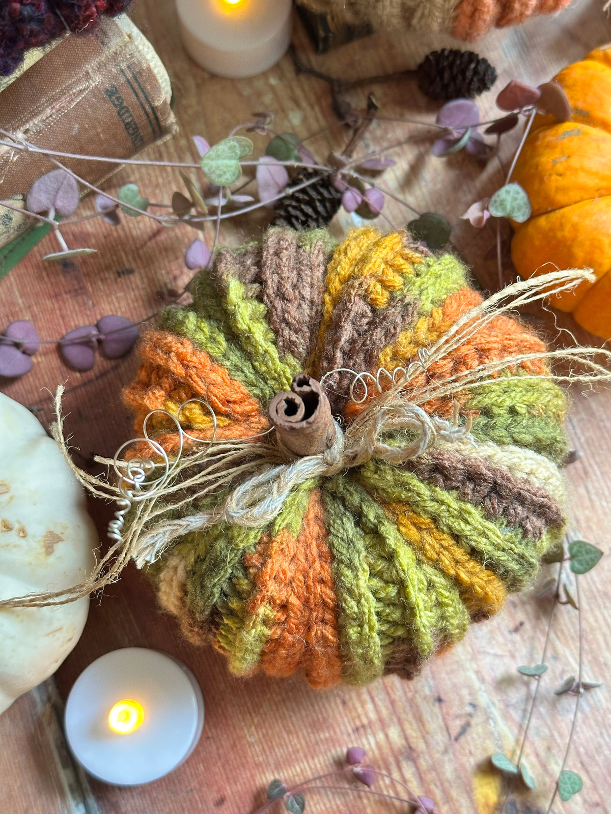 A crocheted autumn pumpkin decoration in warm orange, olive green, mustard yellow, and brown yarn, styled with twine and a cinnamon stick on a rustic wooden surface with candles and seasonal decor.