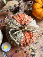 A crocheted decorative pumpkin in autumn colours of orange/pink, beige, and grey, tied with a jute twine bow and a cinnamon stick on a rustic wooden surface with autumnal decorations.