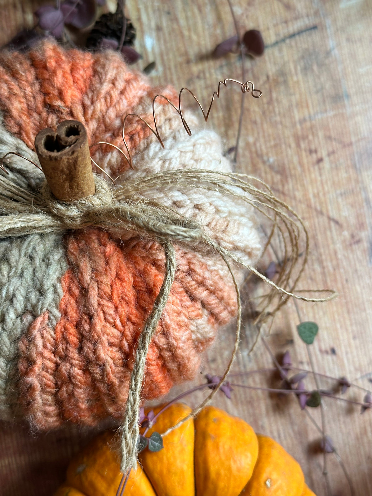 A close-up of a crocheted autumn pumpkin decoration in orange/pink, cream, and beige yarn, tied with twine and topped with a cinnamon stick, set on a wooden surface with a small orange pumpkin below.