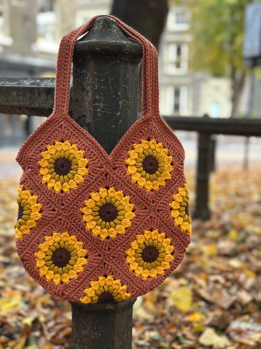 A handmade crocheted shoulder bag with a sunflower pattern, hanging from a dark metal post in an outdoor park setting with fallen leaves.