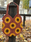 A handmade crocheted shoulder bag with a sunflower pattern, hanging from a dark metal post in an outdoor park setting with fallen leaves.